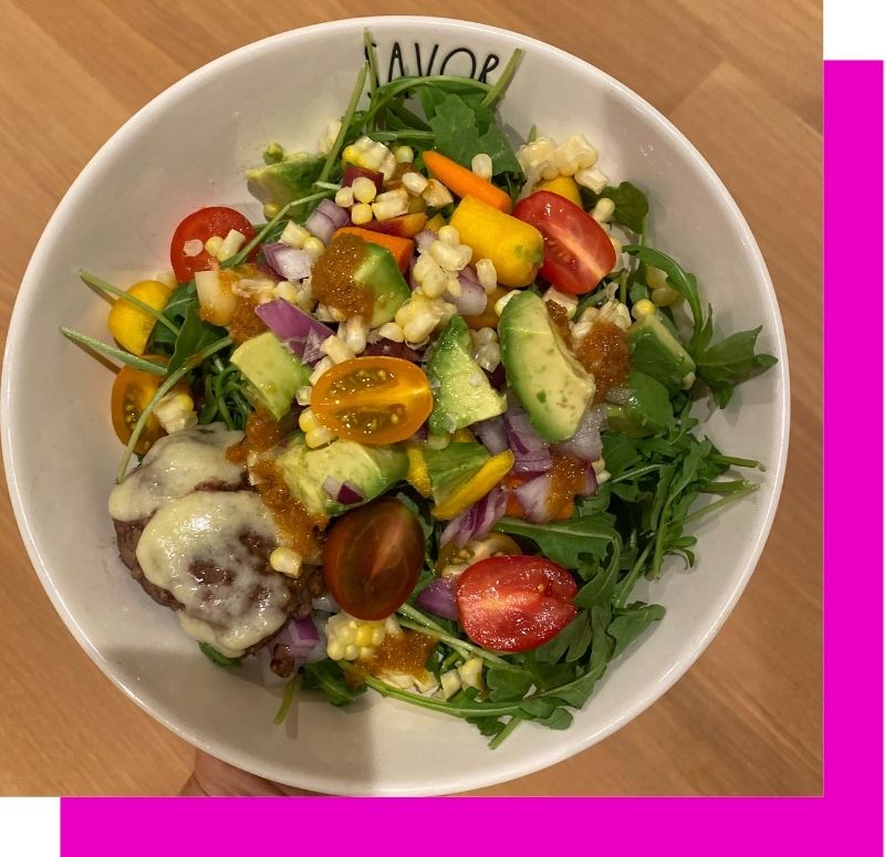 burger bowl of green salad, tomatoes, corn, avocado and red onion topped with a burger